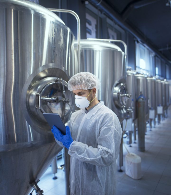 Factory worker technologist in protective uniform with hairnet and mask controlling food production on tablet computer.