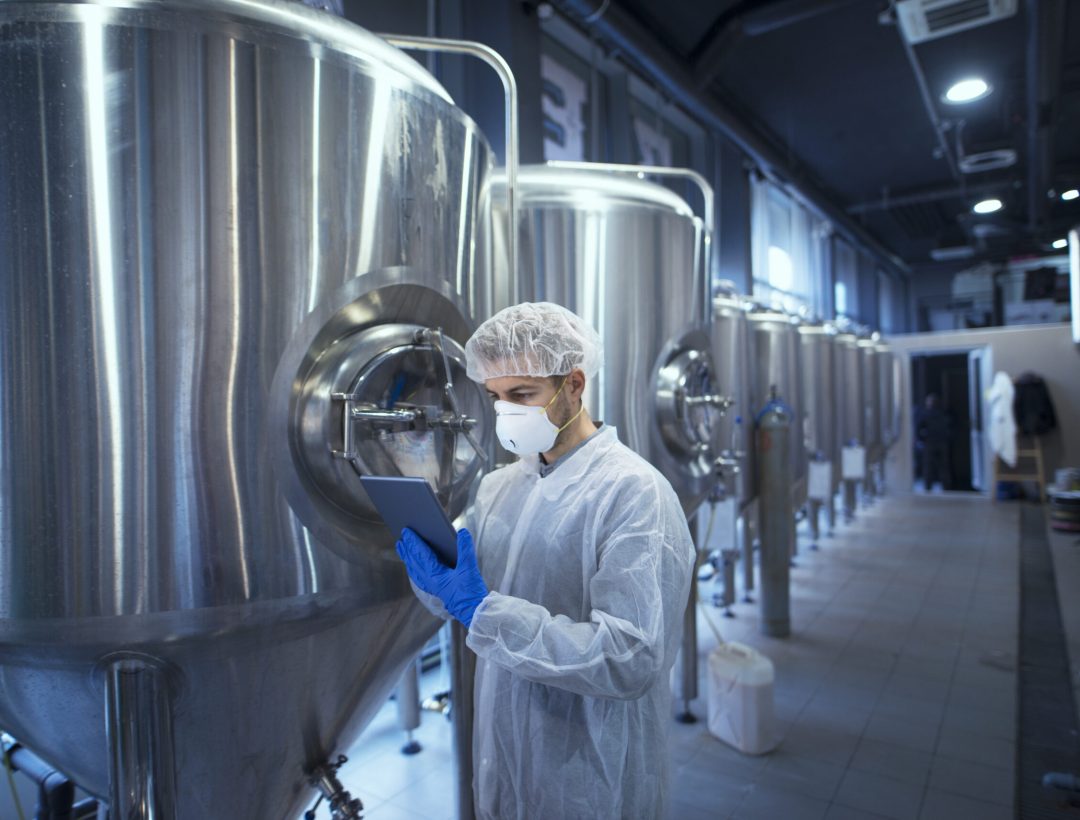 Factory worker technologist in protective uniform with hairnet and mask controlling food production on tablet computer.